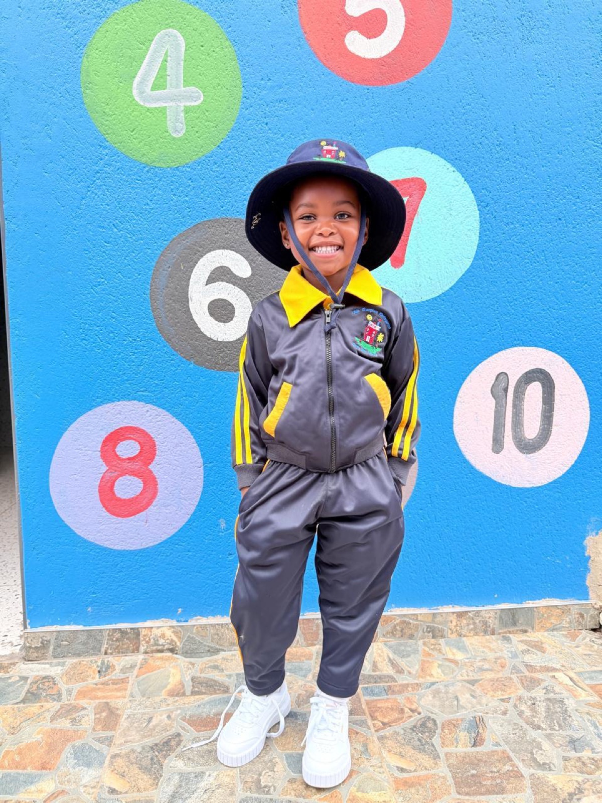A smiling girl in full Little Champions tracksuit uniform and hat at the numbers wall