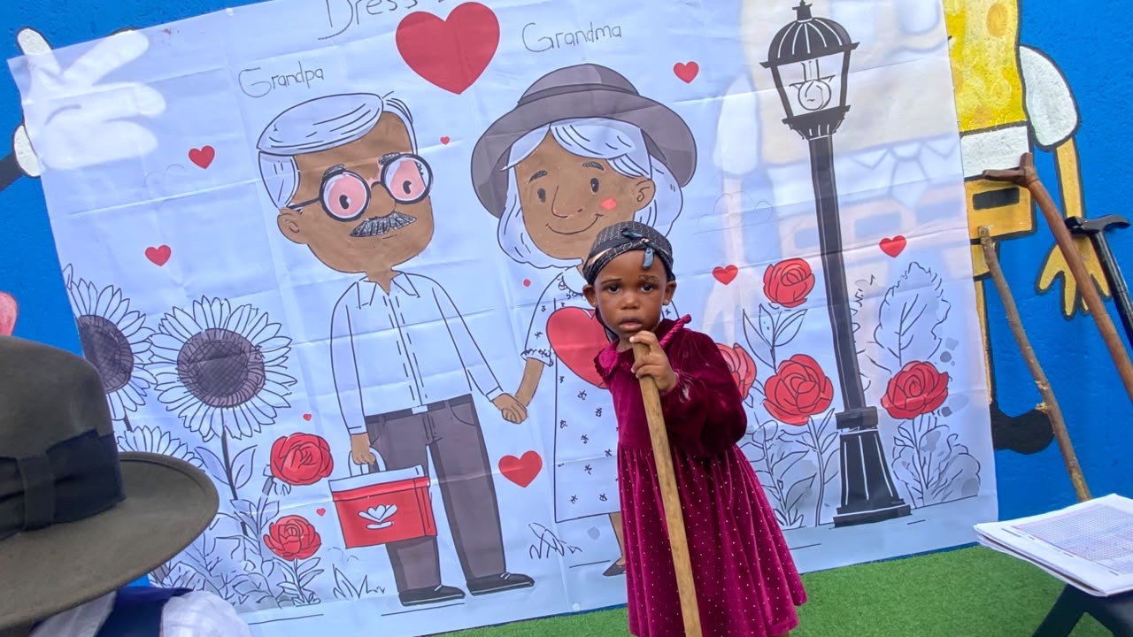 A girl in a velvet dress and headwrap with a walking stick at the Grandparents' Day banner